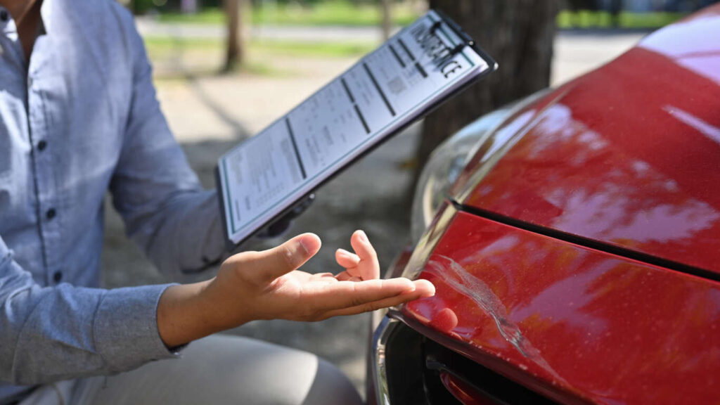 Person holding an insurance document while examining a damaged red car, illustrating the process of navigating insurance requirements after a car accident in North Carolina.