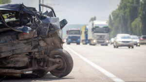 The crushed front end of a black car is shown on the shoulder of a highway, with large trucks and other vehicles visible in the background. This image represents the devastating multi-vehicle crash on I-485 in West Charlotte that resulted in six fatalities.