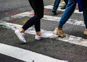 A low-angle shot of a group of people's legs and feet as they walk across a crosswalk on a city street, representing pedestrians in an urban environment.