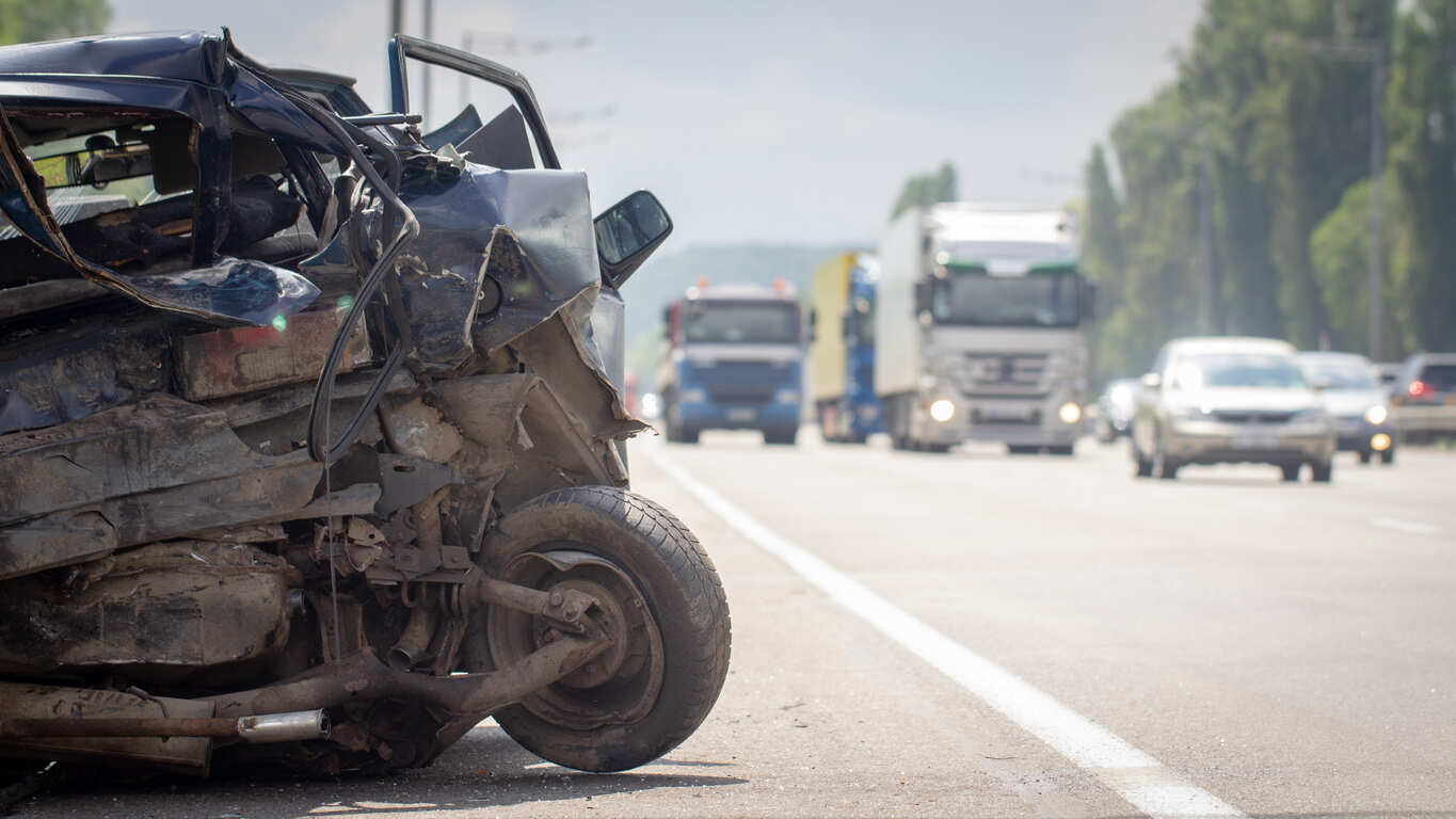 The crushed front end of a black car is shown on the shoulder of a highway, with large trucks and other vehicles visible in the background. This image represents the devastating multi-vehicle crash on I-485 in West Charlotte that resulted in six fatalities.