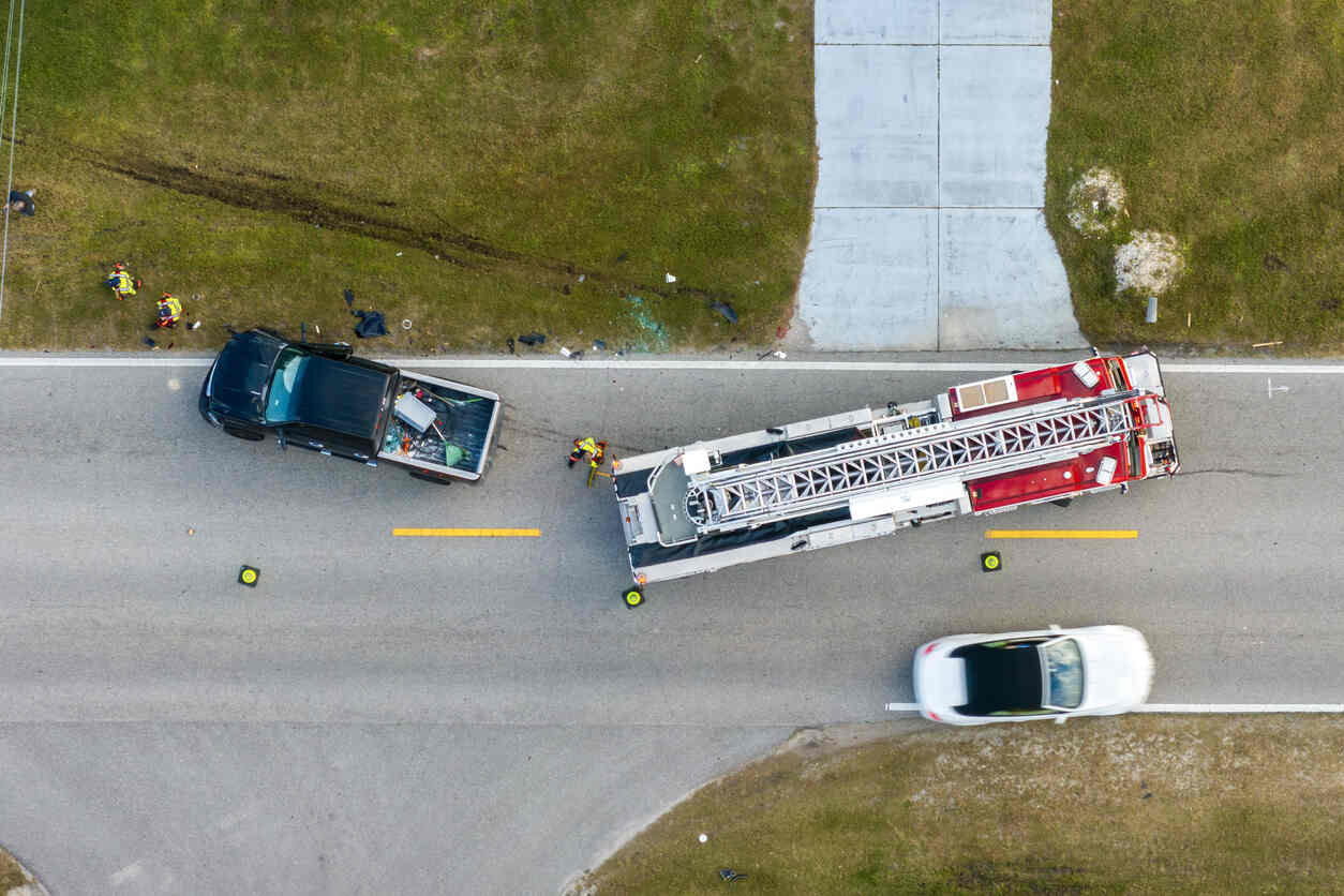 An aerial view of a two-vehicle crash site on a road, with a black pickup truck and a white car. A fire truck is also on the scene.